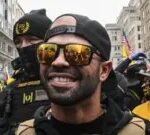 Getty Images Former Proud Boys leader Enrique Tarrio is shown smiling while wearing mirrored sunglasses and back and yellow protective clothing. He is standing outside Harry