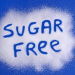 A blue background sprinkled with white sugar substitute crystals with the words sugar free written; concept is sugar alcohols and artificial sweeteners