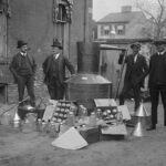 Onlookers watch as suited men stand in front of a large copper kettle still for making illegal liquor with boxes of bottles and funnels spread before them all for the manufacture of booze.