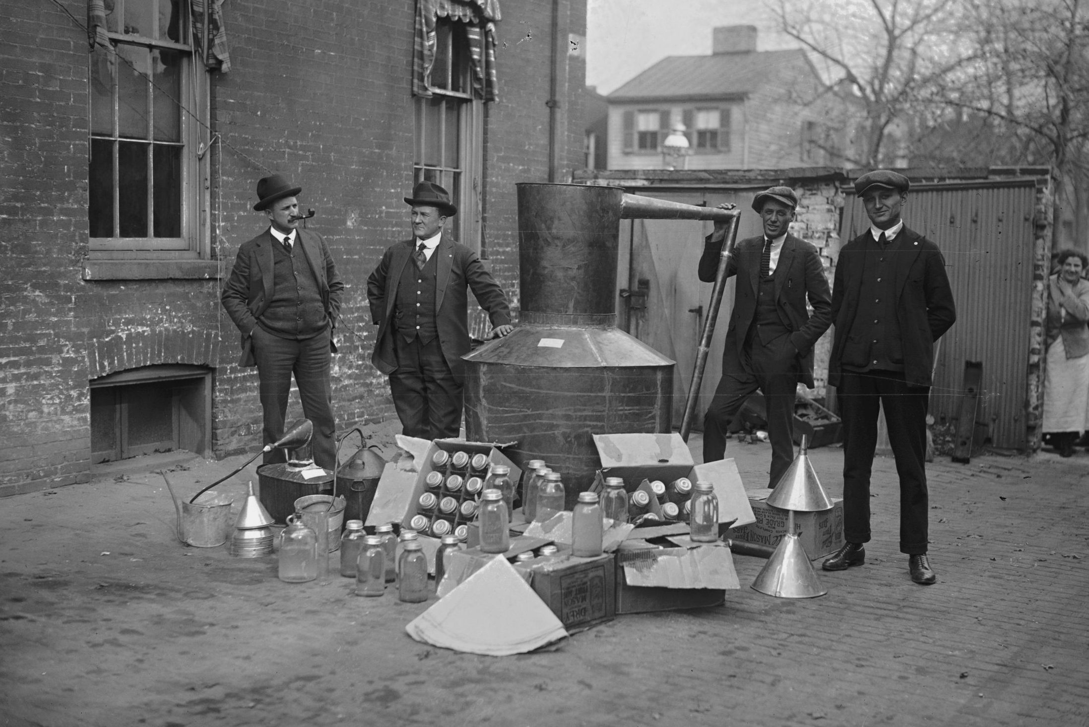 Onlookers watch as suited men stand in front of a large copper kettle still for making illegal liquor with boxes of bottles and funnels spread before them all for the manufacture of booze.