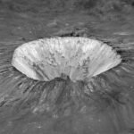 Pierazzo Crater is seen obliquely. Slopes rising to form the wall of the crater are clear. The interior of the crater is bright white mottled with gray. The landscape looks to be flowing outward from the interior of the crater and over its walls.