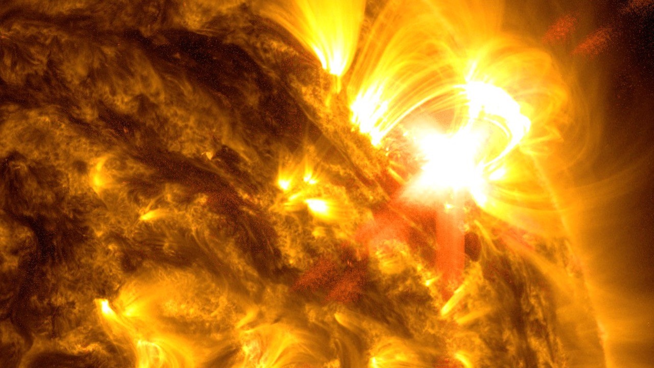 A close-up view shows a portion of the Sun in shades of yellow and gold. A bright, yellow-white flare appears on the right edge of the Sun, illustrating a solar flare event.