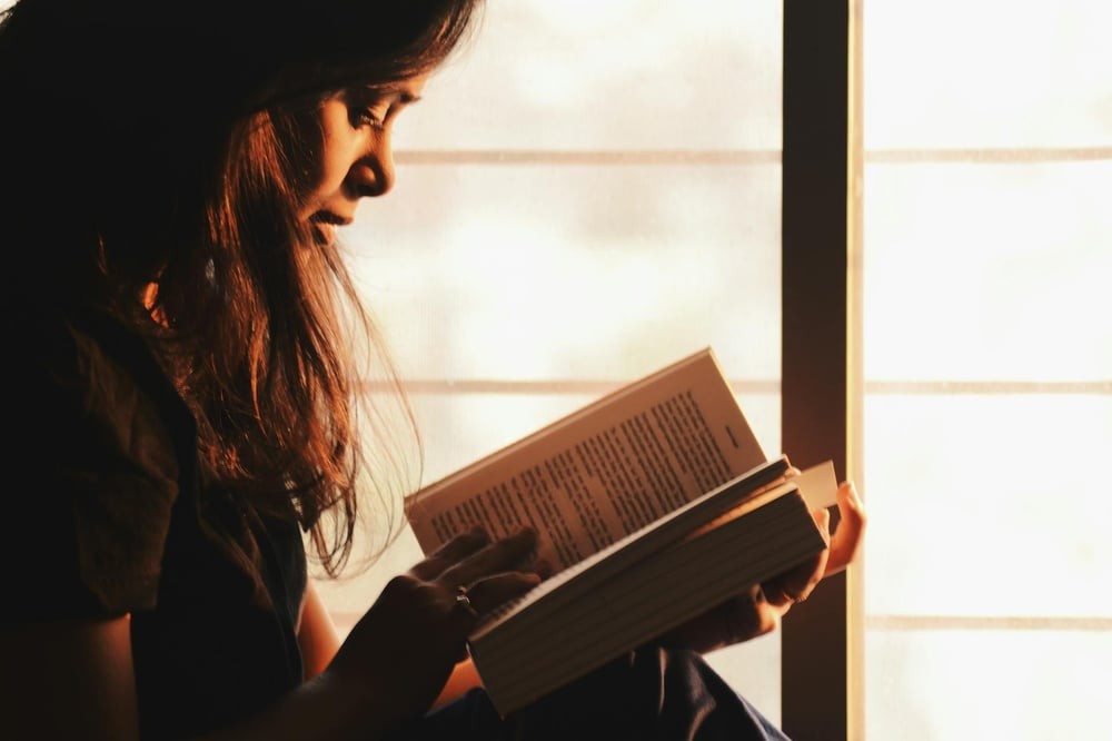 A person reads a book beside a window in a dark room.