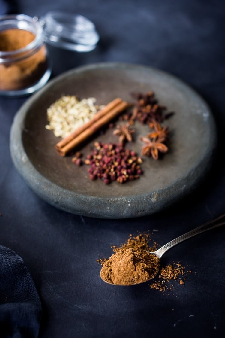Spoonful of homemade Chinese Five Spice in front of dish with whole spices used to make it: Szechuan peppercorns, star anise, cloves, cinnamon stick, fennel seeds