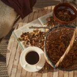 photo viewed from above of a cup of coffee on a saucer next to a basket and a bowl each holding freshly picked mushrooms; everything is on a small round wood table with additional loose mushrooms spread on an open magazine