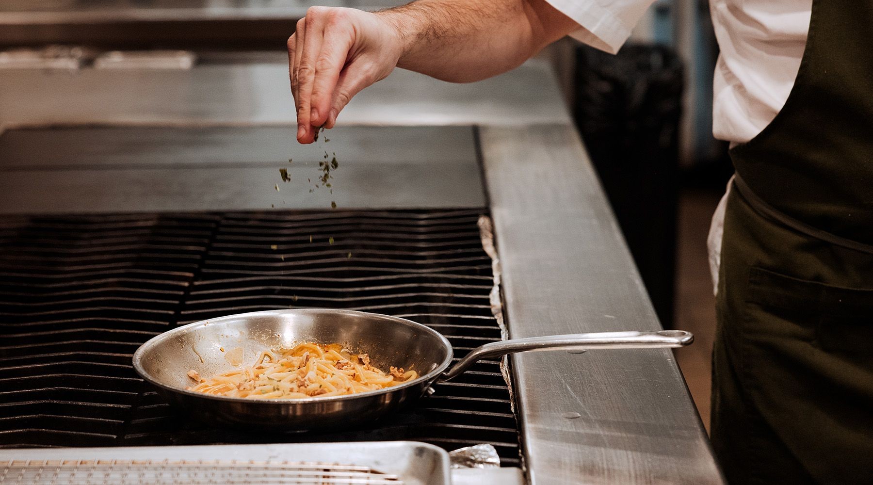 A chef seasons food in a pan on a stove, alt text is The culinary artistry of a chef seasoning food in a skillet on a professional stovetop.