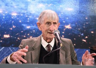 A man speaks into a microphone and gestures with his hands, there is a blue and black space themed background behind.