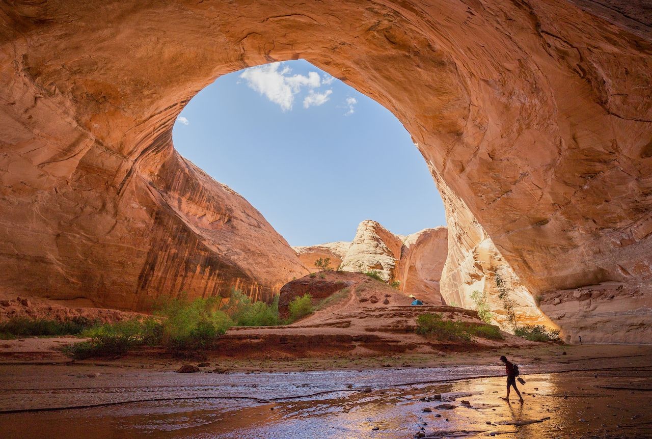 A hiker traversing Jacob Hamblin Arch in Grand Staircase-Escalante National Monument, Utah. The alt text is: A male hiker with a backpack walks through Jacob Hamblin Arch in Coyote Gulch, Grand Staircase-Escalante National Monument, Utah, captured in a wide-angle shot.