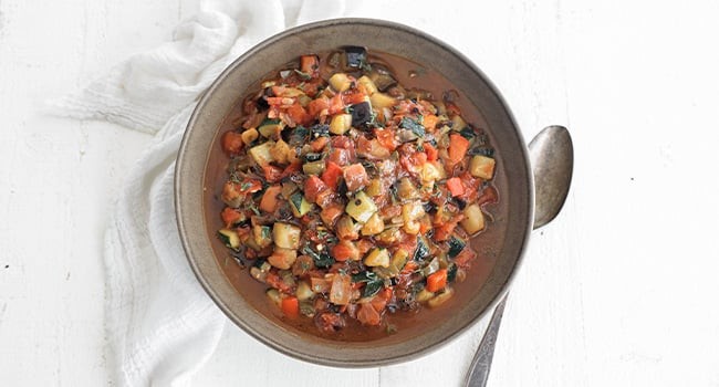 Close-up of ratatouille with a spoon, highlighting the texture and colorful mix of vegetables, garnished with fresh thyme leaves.