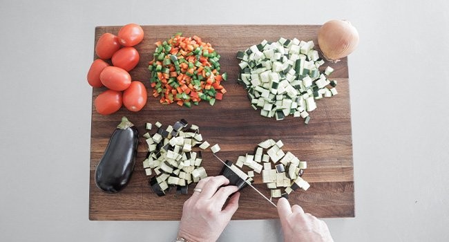 Preparing vegetables for ratatouille, dicing eggplant, zucchini, bell peppers, onions, and tomatoes into evenly sized pieces for consistent cooking.