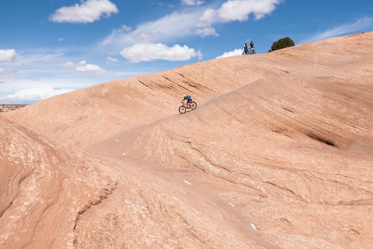 Mountain bikers conquering the Slickrock Trail in Moab, Utah. The alt text is: Two mountain bikers ride across the red sandstone of the Slickrock Trail in Moab, Utah, with the vast desert landscape stretching out behind them.