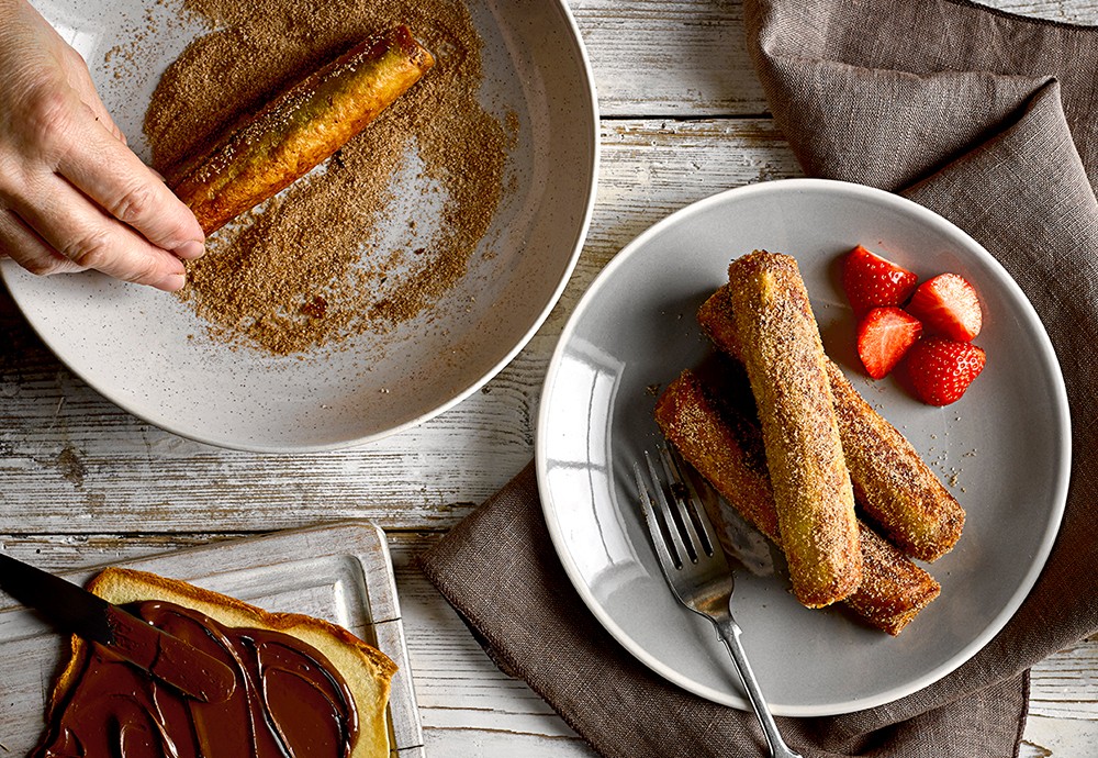 Slices of brioche loaf rolled up and dusted in a cinnamon sugar on two plates with chopped strawberries for a Nutella Roll-Up