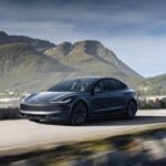 A grey Tesla Model 3 drives along a coastal road