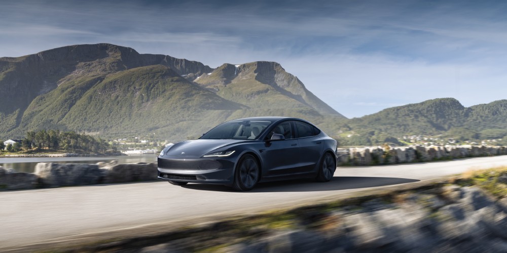 A grey Tesla Model 3 drives along a coastal road