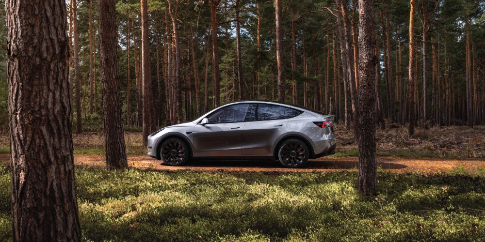 A grey Tesla Model Y car cruises down a road surrounded by trees